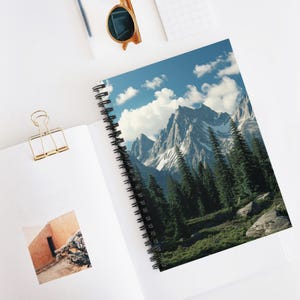 May include: Spiral-bound notebook displaying a mountain landscape with snow-covered peaks, evergreen trees, and a blue sky with white clouds. The notebook is open on a white surface.
