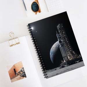 May include: Spiral-bound notebook featuring a space-themed design. The cover displays a detailed illustration of a rocket on a lunar surface, with Earth and the moon visible in the background. The notebook has a black spiral binding.