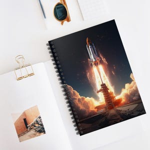 May include: Spiral-bound notebook featuring a vibrant image of a space shuttle launching into space. The cover showcases the shuttle ascending through a fiery sky, with billowing clouds and a launch tower. The notebook is open, revealing blank white pages.
