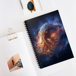Op de afbeelding: Spiraalgebonden notitieboek met een kosmische illustratie van een wezen met gloeiende oranje en gele tentakels tegen een donkerblauwe en paarse achtergrond. Het notitieboek heeft een zwarte spiraalbinding.