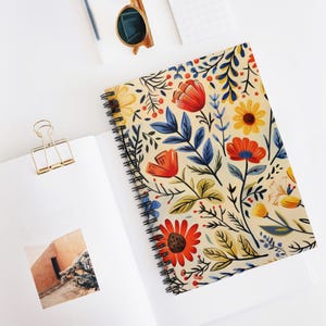Può includere: Quaderno a spirale con motivo floreale. La copertina presenta uno sfondo color crema con fiori e foglie rosse, arancioni, gialle e blu. Il quaderno è aperto su una pagina bianca con una clip dorata.