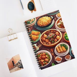 Puede incluir: Cuaderno de espiral con una colorida ilustración de varios platos asiáticos, incluyendo fideos, empanadillas y rollitos de primavera. El cuaderno está abierto sobre una superficie blanca, con un clip dorado y gafas de sol.
