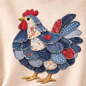 May include: A beige long-sleeved shirt with an appliqué of a rooster. The rooster is made from denim, floral, and polka dot fabrics, with a red comb and heart. The rooster's legs are a golden yellow colour.