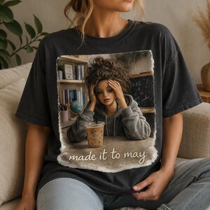 May include: Dark gray t-shirt featuring a distressed graphic of a person with their head in their hands, a drink, and the text "made it to may". The graphic includes a bookshelf, globe, and chalkboard.