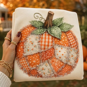 May include: A cream-colored sweatshirt featuring a large, central pumpkin applique made from various orange and white fabric patches with frayed edges. The pumpkin has a textured brown stem and green fabric leaves at the top.