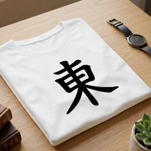 Puede incluir: Camiseta blanca con un dise&ntilde;o de car&aacute;cter Kanji japon&eacute;s negro. La camiseta est&aacute; doblada sobre una superficie de madera, con un reloj y una peque&ntilde;a planta visibles en el fondo.