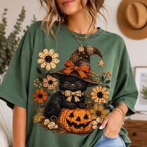 Puede incluir: Una camiseta verde oliva con un estampado gr&aacute;fico de un gato negro con un sombrero de bruja a cuadros y una pajarita a juego. El gato est&aacute; sentado sobre una calabaza naranja tallada rodeada de flores y follaje amarillo y naranja.