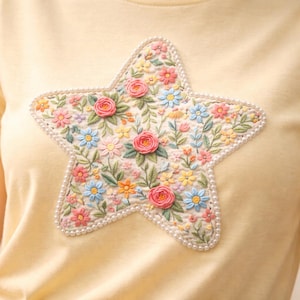 Może przedstawiać: Jasnożółta koszulka z haftem kwiatowym w kształcie gwiazdy. Haft zawiera różowe róże, niebieskie i żółte kwiaty oraz zielone liście, obrysowane małymi białymi koralikami. Koszulka wykonana jest z miękkiej, jasnej tkaniny.