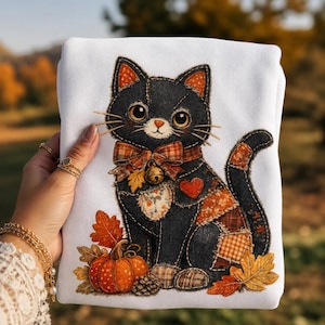 Puede incluir: Una sudadera blanca con una gran aplicación detallada de un gato negro con detalles de tela estilo patchwork en cuadros naranjas y marrones. El gato lleva un lazo con un cascabel y está rodeado de calabazas naranjas y hojas de otoño.