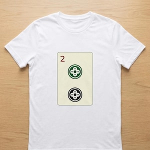 Puede incluir: Camiseta blanca con un dise&ntilde;o de carta de juego. La carta tiene un fondo beige con el n&uacute;mero "2" en rojo en la parte superior. Debajo, dos s&iacute;mbolos estilizados en forma de tr&eacute;bol, uno verde y otro negro.