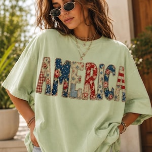 Könnte beinhalten: Ein hellgrünes Kurzarm-T-Shirt mit dem Wort AMERICA auf der Brust. Jeder Buchstabe ist mit einem anderen Patchwork-Muster gefüllt, darunter rot-weißes Vichy-Karo, blaue Blumen, rote Blumen sowie rot-weiße Streifen.