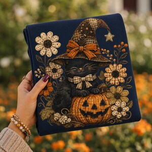 Puede incluir: Una tela azul oscuro con un estampado gr&aacute;fico de un gato negro con un sombrero de bruja de parches marrones y una pajarita. El gato est&aacute; sentado sobre una calabaza naranja tallada rodeada de flores marrones y crema y estrellas.
