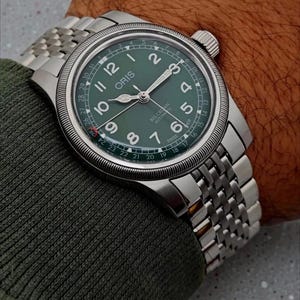 May include: A silver wristwatch with a green face and a stainless steel band. The watch face displays the brand name "ORIS" and the words "BIG CROWN AUTOMATIC". The watch has a date display and luminous hands. The watch is on a wrist.