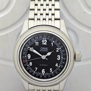 Może przedstawiać: Srebrny zegarek na rękę z czarną tarczą. Zegarek posiada wyświetlacz daty, świecące wskazówki i bransoletę ze stali nierdzewnej. Na tarczy widnieje nazwa marki "ORIS" oraz napisy "AUTOMATIC" i "25 JEWELS".