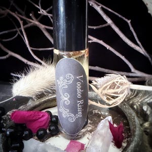 May include: A clear glass roll-on bottle with a black cap, labeled "Voodoo Rising" in white script. The bottle is surrounded by dried flowers, crystals, and black beads. The background is dark, with branches and a feather.