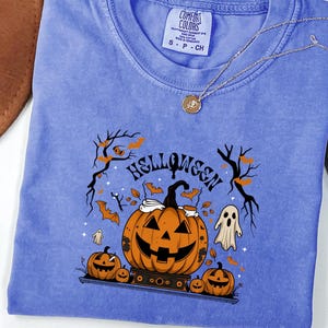 May include: A periwinkle blue short-sleeve t-shirt featuring a graphic print of three jack-o'-lanterns, a ghost, and bare tree branches. The text "HELLOWEEN" is printed above the central pumpkin. The shirt has a Comfort Colors brand label.