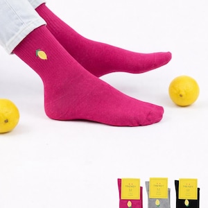 Könnte beinhalten: Leuchtend pinke Socken mit einer kleinen Zitronenstickerei am Knöchel. Die Socken sind gerippt und scheinen aus einem weichen Material zu sein. Zwei Zitronen liegen in der Nähe der Füße. Drei Paar Socken in Pink, Grau und Schwarz werden mit gelben Etiketten gezeigt.