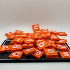 May include: Orange letter tiles with white letters are scattered on a black tray. The letters are in a variety of fonts and sizes. The background is white.