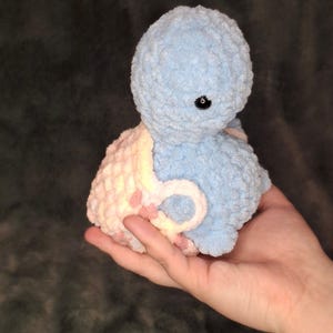 May include: A handmade blue and pink crochet octopus toy with a single black eye. The plushie is held in a hand, showcasing its soft, textured yarn. The octopus has a round head and a curved body.