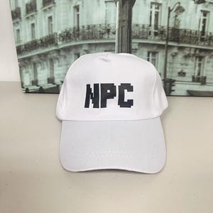 Puede incluir: Gorra de béisbol blanca con las letras negras "NPC" en una fuente pixelada. La gorra tiene una visera curva y una corona estructurada. El fondo presenta una imagen en blanco y negro de una escena callejera.