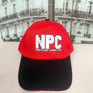 Puede incluir: Gorra de béisbol roja y negra con las letras blancas "NPC" en bloques. El texto "NON PLAYER CHARACTER" está debajo. La gorra tiene una visera negra y un botón negro en la parte superior.
