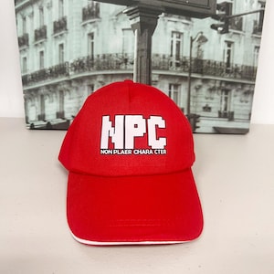 Puede incluir: Gorra de béisbol roja brillante con ribete blanco. La gorra presenta las letras blancas "NPC" sobre el texto "NON PLAYER CHARACTER". El sombrero es un diseño clásico de seis paneles.