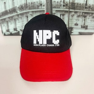 Puede incluir: Gorra de béisbol negra y roja con el texto blanco "NPC NON PLAYER CHARACTER". La gorra tiene una visera roja y un botón rojo. El fondo es una imagen borrosa de un edificio.