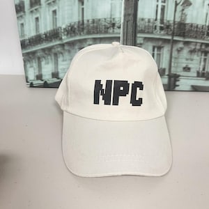 Puede incluir: Gorra de béisbol blanca con visera curva. La gorra presenta el acrónimo "NPC" en letras negras en la parte delantera. El sombrero está hecho de una tela suave y es adecuado para ropa casual.
