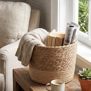 May include: A round, woven jute basket with a thick rope texture, containing a beige cable-knit throw blanket, several hardback books, and a magazine. The basket sits on a wooden surface alongside a ceramic mug, a succulent, and open spectacles.