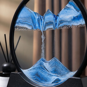 May include: A circular sand art piece with blue and white sand, forming a mountain scene. The black frame and base provide a contrast to the flowing sand art. A decorative item for the home, it adds a touch of artistic flair.