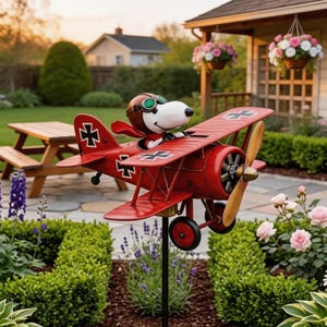 May include: A red metal airplane yard decoration featuring Snoopy as the pilot, wearing a brown aviator helmet and goggles. The biplane has a wooden propeller and black and white cross symbols on the wings. The decoration is set in a garden with flowers and a picnic table.