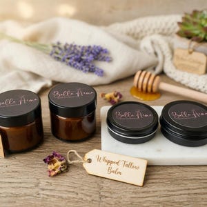 以下が含まれることがあります： 木製の表面に、ホイップタローバームの瓶や缶など、さまざまな美容製品が並んでいます。「Belle Aima」の文字が入った黒い蓋とラベルが付いています。ラベンダーの小枝と多肉植物も含まれています。