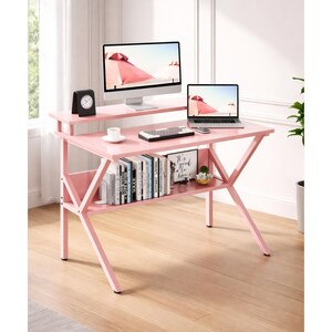 May include: A pink computer desk with a top shelf, lower shelf, and angled legs. A computer monitor, laptop, keyboard, and clock are on the desk. Books and a small plant are on the lower shelf. The desk is 47 inches wide.