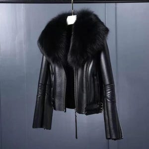 May include: Black leather jacket with a large, fluffy black fur collar. The jacket has a zipper closure, long sleeves with zippers at the cuffs, and decorative stitching on the sleeves. The jacket is hanging from a metal bar.