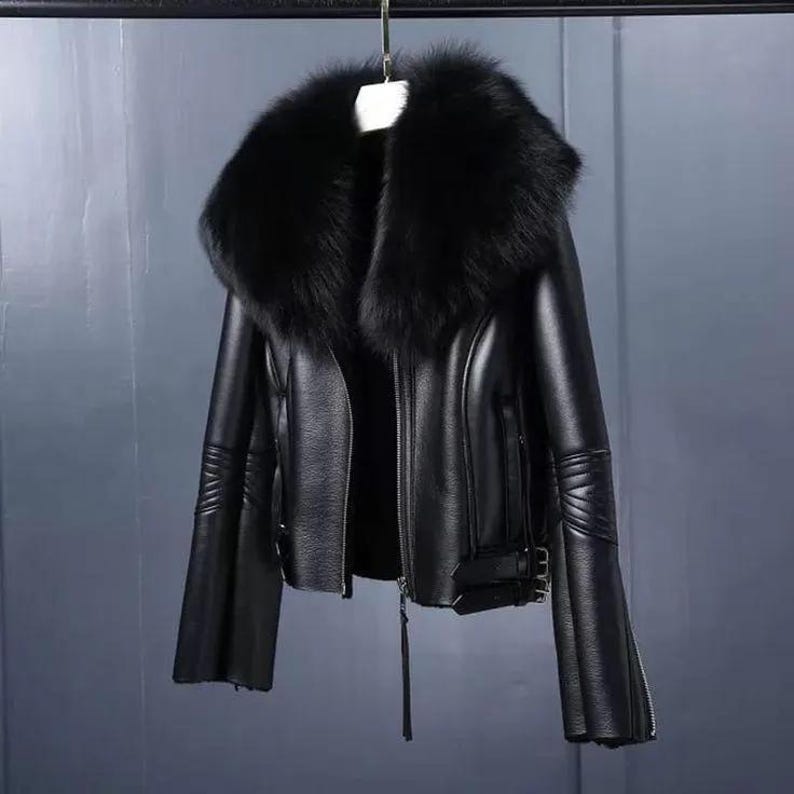 May include: Black leather jacket with a large, fluffy black fur collar. The jacket has a zipper closure, long sleeves with zippers at the cuffs, and decorative stitching on the sleeves. The jacket is hanging from a metal bar.