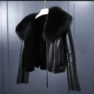 May include: A black leather jacket with a large, fluffy black fur collar. The jacket has a zipper closure, a belt buckle detail, and zippered sleeves. The jacket is hanging on a white hanger against a gray background.