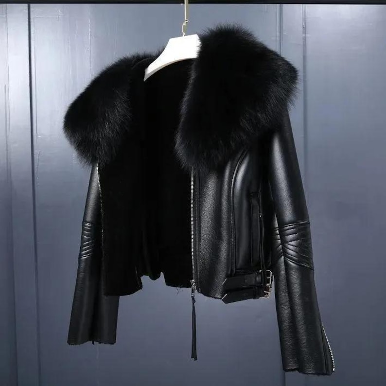 May include: A black leather jacket with a large, fluffy black fur collar. The jacket has a zipper closure, a belt buckle detail, and zippered sleeves. The jacket is hanging on a white hanger against a gray background.