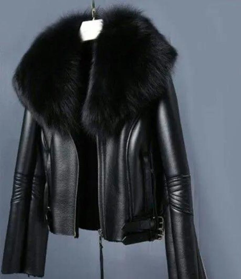 May include: A black leather jacket with a large, fluffy black fur collar. The jacket has a zipper closure, flared sleeves, and buckle details. The jacket is hanging on a hanger.