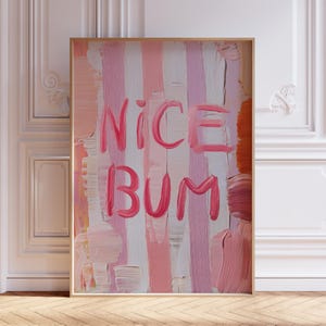 Op de afbeelding: Een ingelijst kunstwerk met een gouden rand. Het schilderij toont verticale penseelstreken in tinten roze, wit en oranje. Vetgedrukte, roze tekst luidt "NICE BUM" over het midden van het doek. Het kunstwerk wordt tentoongesteld tegen een witte muur.