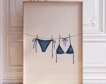 May include: A framed watercolor print featuring a dark blue bikini set hanging from a thin string with wooden clothespins. The bikini consists of a triangle top and side-tie bottoms, rendered in a minimalist style against a light beige background.