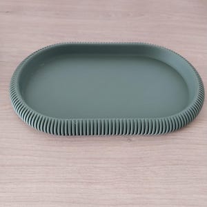 May include: A sage green oval tray with a ribbed border. The tray features a smooth, flat interior and a raised, textured edge. The design is simple and modern, suitable for various decorative or functional uses.