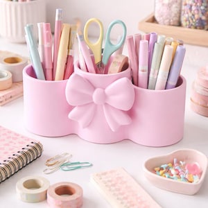 May include: A pink desk tidy with a bow design, holding pastel pens, pencils, and scissors. Washi tape, paper clips, and a heart-shaped dish are also on display, creating a coordinated stationery set.