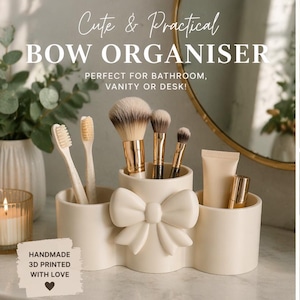 May include: A cream-coloured bow-shaped organiser with three compartments, holding toothbrushes, makeup brushes, and beauty products. The text reads "Cute & Practical BOW ORGANISER" and "HANDMADE 3D PRINTED WITH LOVE".