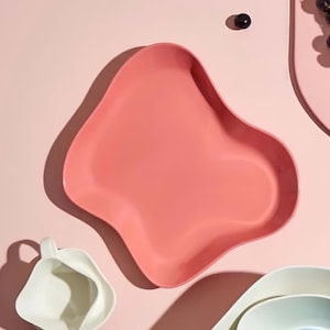 Może przedstawiać: Kolekcja kolorowych, organicznych kształtów naczyń do serwowania w odcieniach różu, błękitu i bieli. Niektóre naczynia zawierają winogrona i kumkwaty. Układ znajduje się na różowym tle, tworząc nowoczesną i stylową estetykę.