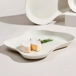 Może przedstawiać: Biały ceramiczny talerz do serwowania o unikalnym, falistym kształcie. Na talerzu ułożono kawałek sera brie i gałązkę rozmarynu. Dodatkowe białe naczynia do serwowania są widoczne w tle. Obraz jest dobrze oświetlony, podkreślając faktury.