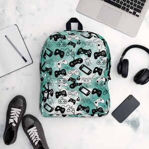 May include: A teal blue backpack with a white and black pattern of video game controllers. The backpack has a zipper closure and adjustable straps.