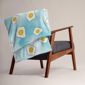 May include: A blue and white throw blanket with a pattern of yellow fried eggs. The blanket is draped over a wooden chair with a gray cushion.