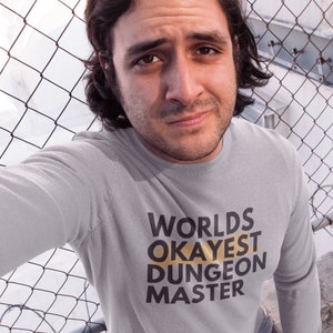 May include: A gray long-sleeve shirt with the text "Worlds Okayest Dungeon Master" printed in black and gold letters.