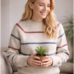 Pode incluir: Um suéter creme com listras vermelhas e azuis. O suéter tem gola redonda e mangas compridas. Uma pequena planta em vaso é segurada em frente ao suéter.