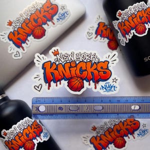 May include: A die-cut sticker featuring the text New York Knicks in orange and blue graffiti-style lettering with a basketball graphic, a crown, and hearts. A blue ruler is placed across the sticker, showing a width of approximately 5.5 inches.